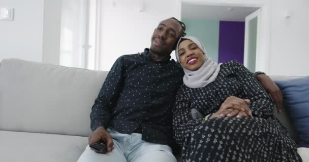 Αφρικανικό ζευγάρι κάθεται στον καναπέ βλέποντας τηλεόραση μαζί γυναίκα φορώντας ισλαμικά ρούχα μαντίλα — Αρχείο Βίντεο