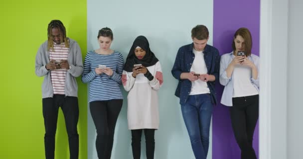 Багатокультурна група друзів за допомогою мобільних телефонів Студенти стоять в рядку і друкують на смартфонах над кольоровою стіною — стокове відео