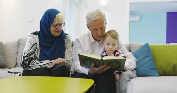 Kakek-nenek keluarga muslim tradisional dengan anak-anak membaca Al-Quran dan berdoa bersama di sofa — Stok Video