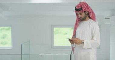 Genç ve modern Müslüman Arap iş adamı geleneksel kıyafetler giyerken akıllı telefon kullanıyor.
