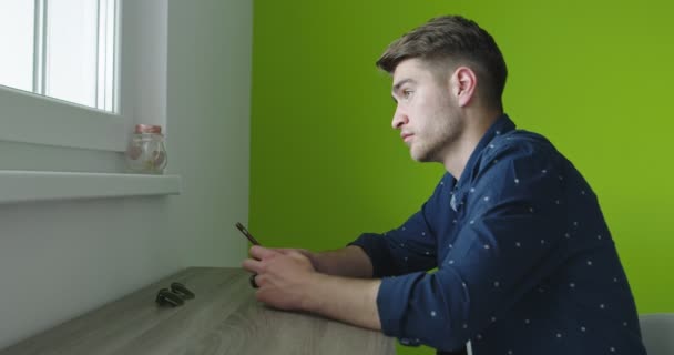 Усміхнений красивий молодий чоловік, використовуючи телефон біля вікна над зеленою стіною — стокове відео