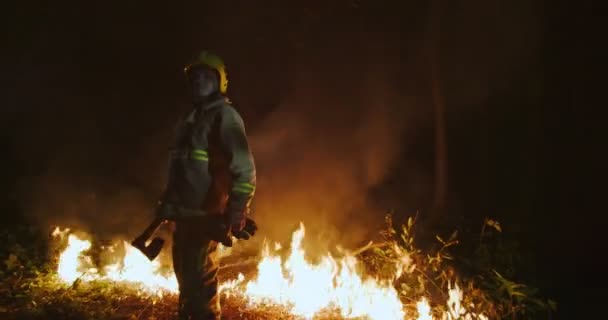 A tűzoltó teljes felszereléssel a kezében tartja a baltát, és egy lángoló erdő hátterében áll. — Stock videók