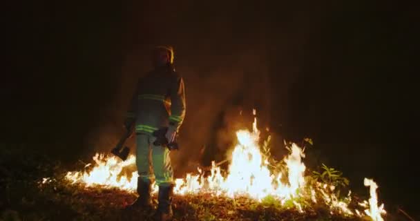 Hasič s plnou výstrojí, drží sekeru v ruce, stojí na pozadí lesa v plamenech — Stock video