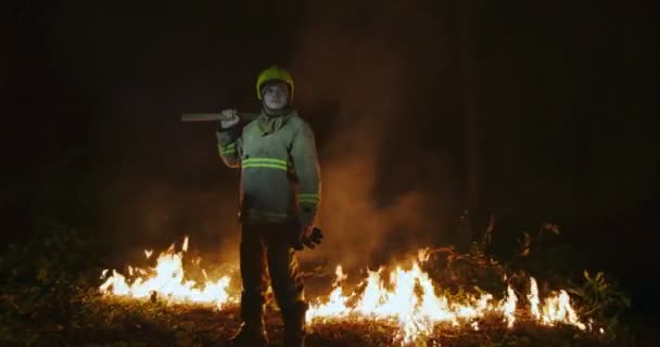 Brandman med full utrustning, håller yxan i handen, står mot bakgrund av en skog i lågor — Stockvideo