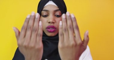 Genç ve güzel Afro-Amerikalı kız izole edilmiş sarı arka planda Müslüman başörtüsü takıyor ve el ele dua ediyor.