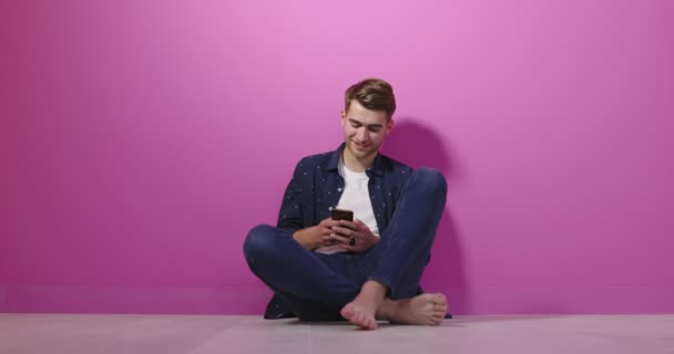 Усміхнений молодий чоловік тримає смартфон і дивиться на нього. Портрет щасливого чоловіка з використанням мобільного телефону ізольовано на рожевому фоні . — стокове відео