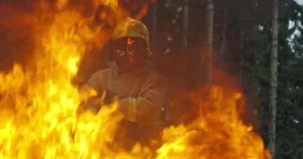 A tűzoltó teljes felszereléssel a kezében tartja a baltát, és egy lángoló erdő hátterében áll. — Stock videók