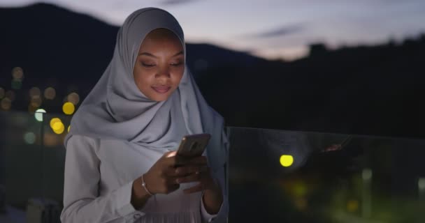 Jovem muçulmana vestindo véu de cachecol na rua urbana da cidade à noite mensagens de texto no smartphone com luz da cidade bokeh no fundo — Vídeo de Stock