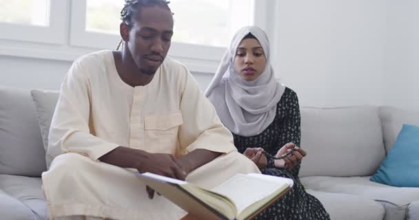 Αφρικανικό μουσουλμανικό ζευγάρι στο σπίτι στο Ραμαζάνι ανάγνωση Quran Holly Islam βιβλίο στο σύγχρονο διαμέρισμα στον καναπέ — Αρχείο Βίντεο