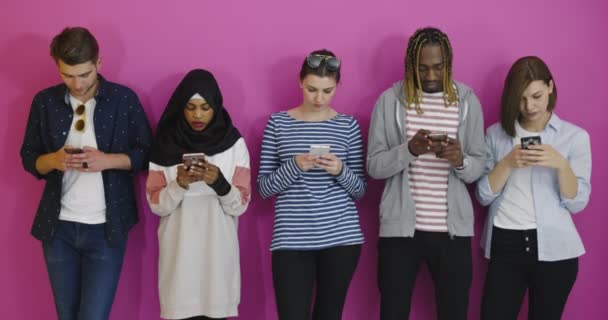 Grupo multicultural de amigos usando teléfonos celulares: estudiantes de pie en fila y escribiendo en los teléfonos inteligentes sobre la pared de color — Vídeo de stock
