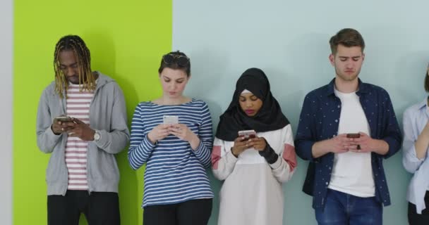 Grupo multicultural de amigos usando teléfonos celulares: estudiantes de pie en fila y escribiendo en los teléfonos inteligentes sobre la pared de color — Vídeos de Stock