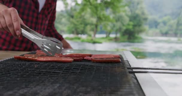 Gros plan de l'homme cuisinant de la viande sur le barbecue à l'extérieur dans la nature au bord de la rivière et préparant pour le dîner dans la nature — Video