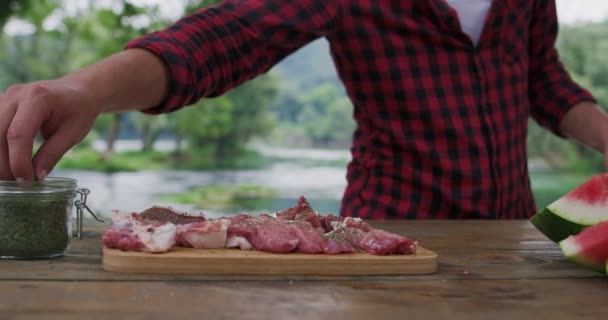 Man preparing meat and puting spices for barbeque and dinner beside river, preparing for dinner in nature in summer kitchen — Stock Video