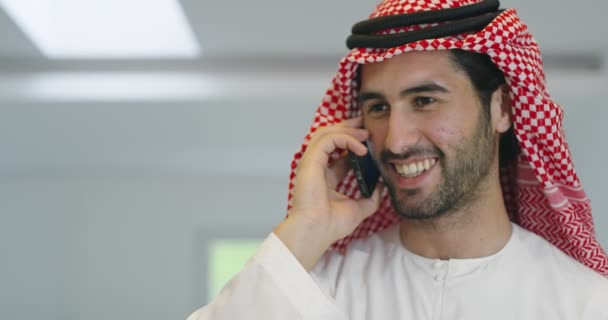 Молодой современный мусульманский арабский бизнесмен в традиционной одежде, используя смартфон дома — стоковое видео