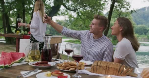 Молода щаслива пара приймає селфі під час пікніка французька вечірка на відкритому повітрі під час літніх канікул біля річки в красивій природі — стокове відео