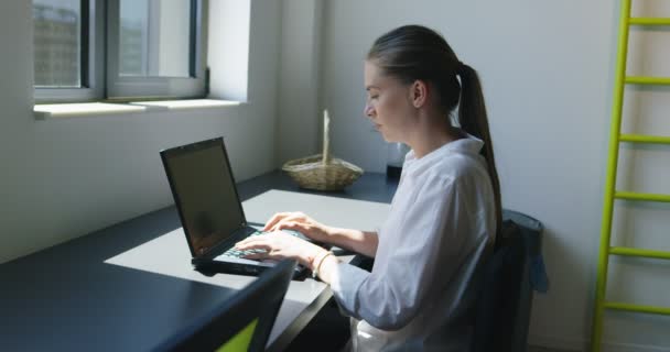 Atractiva joven empresaria alegre trabajando en el ordenador portátil y sonriendo mientras está sentada en su escritorio en la oficina moderna y luminosa — Vídeos de Stock