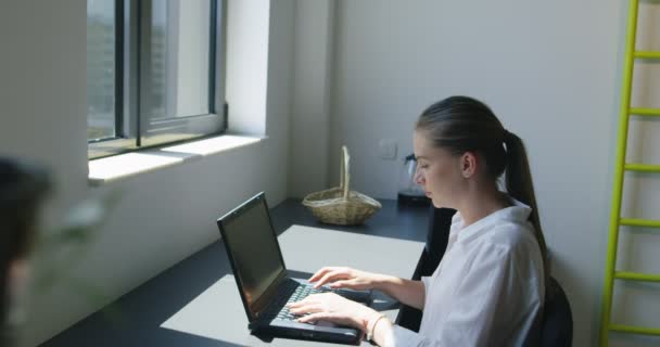 Attraktive, fröhliche junge Geschäftsfrau, die am Laptop arbeitet und lächelt, während sie an ihrem Schreibtisch im hellen modernen Büro sitzt — Stockvideo