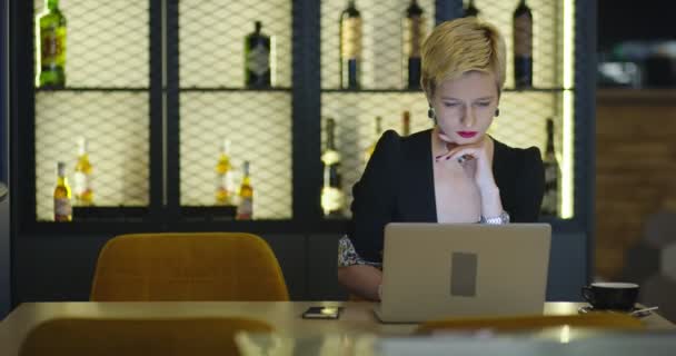 Jovem sentada em uma cafeteria à mesa de madeira, tomando café e usando laptop. Menina navegando na internet, conversando, blogging. — Vídeo de Stock