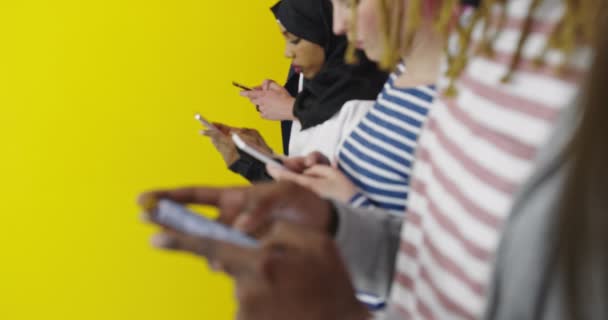 Grupo multicultural de amigos usando teléfonos celulares: estudiantes de pie en fila y escribiendo en los teléfonos inteligentes sobre la pared de color — Vídeos de Stock