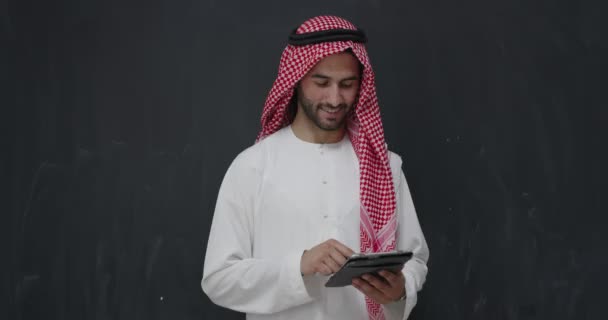 Portrait of young arabian man in traditional clothes in front of black chalkboard rusing tablet — Stock Video