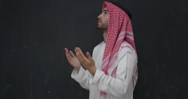 Jonge Arabische man in traditionele kleren maken van traditioneel gebed tot God, houdt de handen in gebed gebaar in de voorkant van zwart schoolbord — Stockvideo