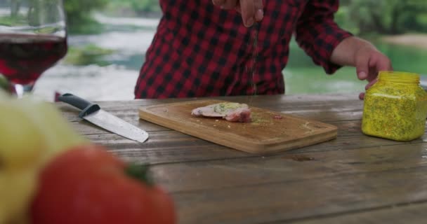 Man preparing meat and puting spices for barbeque and dinner beside river, preparing for dinner in nature in summer kitchen — Stock Video