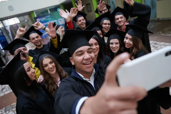 Группа счастливых иностранных студентов в миномётных досках и холостяцких платьях с дипломами, делающими селфи на смартфоне — стоковое фото