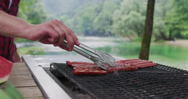 Крупный план человека, готовящего мясо на барбекю на природе рядом с рекой и готовящего ужин на природе — стоковое видео