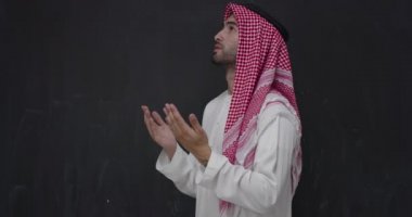 Siyah karatahtanın önünde geleneksel giysiler içinde Müslüman bir adam elleri açık Allah 'a dua ediyor.