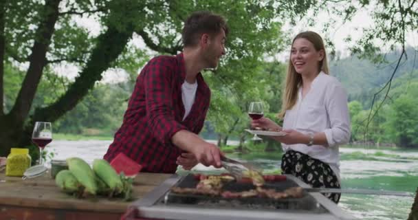 Счастливая пара готовит барбекю и пьет красное вино на открытом воздухе возле реки — стоковое видео