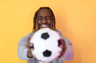 Elinde futbol topu tutarken sarı arka planda poz veren afro bir adam.