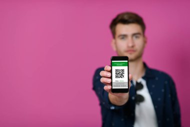 Covid19 pasaport, genç bir adam akıllı telefonunda pembe bir arka plan önünde poz verirken QR kodunu ve Coronavirus pasaportunu gösteriyor.