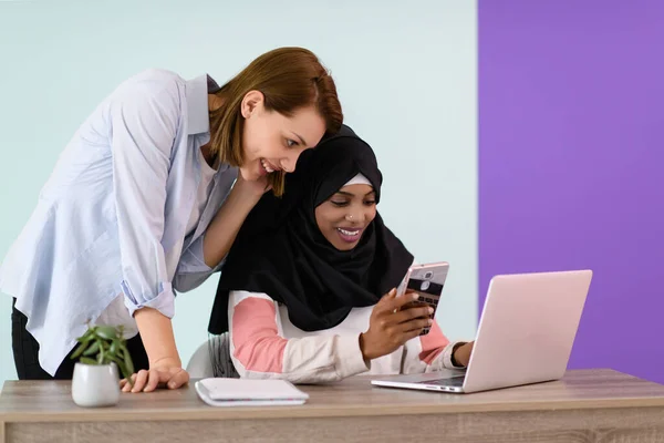 Afro chica con un hijab y una mujer europea utilizar un teléfono celular y portátil en su oficina en casa —  Fotos de Stock