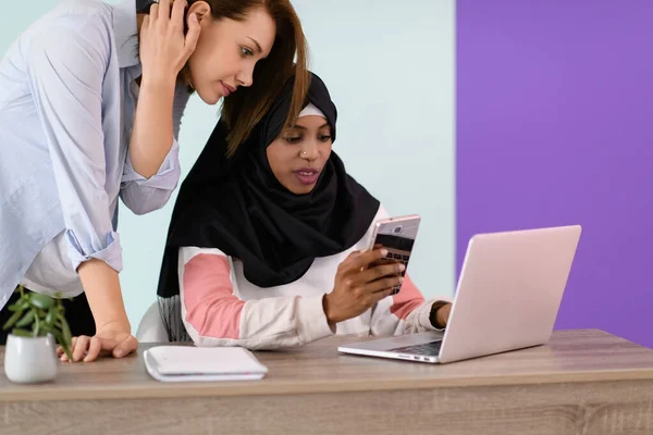 Αφρο κορίτσι με μαντίλα και μια Ευρωπαία χρησιμοποιούν κινητό και λάπτοπ στο γραφείο τους. — Φωτογραφία Αρχείου