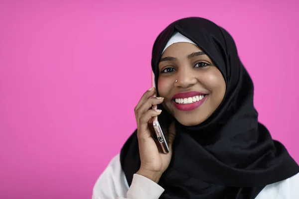 一个面带微笑的漂亮的非洲女孩在粉色背景前广泛地使用手机 — 图库照片