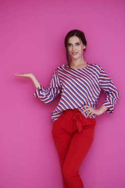 Mutlu gülümseyen genç bir kadının portresi. Pembe arka planda açık avuç içi ile sunumunu yapıyor. Modern modaya uygun giyinmiş, stüdyoda poz veren bir kadın model.