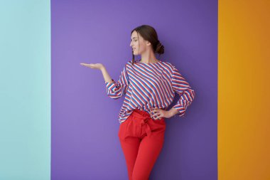 Mutlu gülümseyen genç bir kadının portresi. Renkli arka planda açık avuç içi ile sunum yapan güzel bir kadın. Modern modaya uygun giyinmiş, stüdyoda poz veren bir kadın model.
