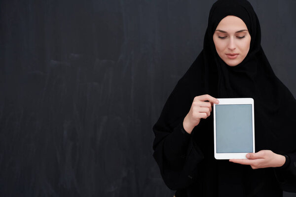 Young Arab businesswoman in traditional clothes or abaya and glasses holding tablet computer in front of black chalkboard representing modern islam fashion and technology