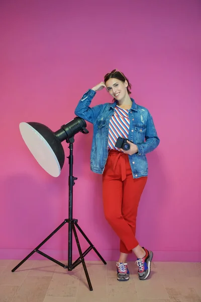 若い女性はスタジオの懐中電灯にもたれてピンクの背景にカメラを持っています スタジオでポーズをとるファッショナブルでカジュアルな服を着た女性写真家 プロの写真 — ストック写真