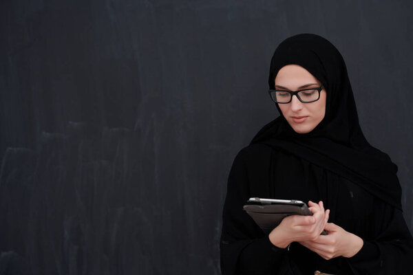 Young Arab businesswoman in traditional clothes or abaya and glasses holding tablet computer in front of black chalkboard representing modern islam fashion and technology