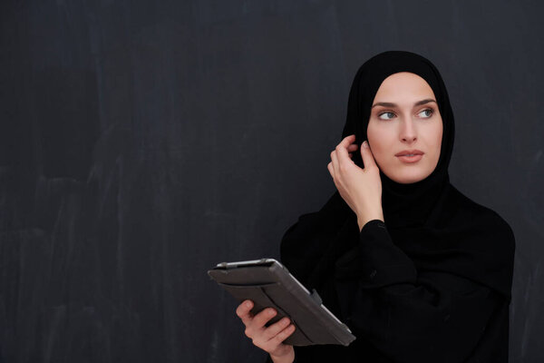 Young Arab businesswoman in traditional clothes or abaya and glasses holding tablet computer in front of black chalkboard representing modern islam fashion and technology