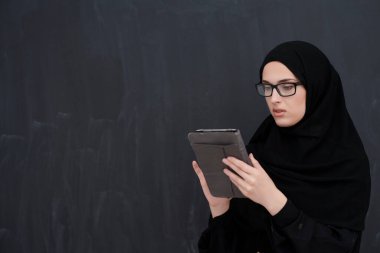 Geleneksel giysiler veya abaya giymiş genç Arap iş kadını ve modern İslami moda ve teknolojiyi temsil eden siyah karatahta tablet bilgisayarın önünde gözlük tutuyor.