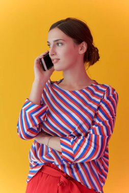 Sarı arka planda dikilirken telefonla konuşan genç kızın portresi. Modern moda ve teknoloji kavramını temsil eden kadın model.