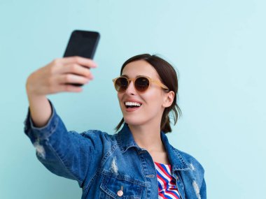 Mavi arka planda selfie çeken genç bir kızın portresi. Güneş gözlüğü takan ve modern moda ve teknolojiyi temsil eden bir kadın model.