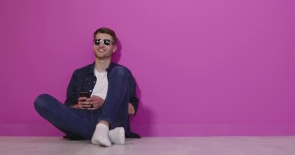 Jovem sorridente segurando o telefone inteligente e olhando para ele. Retrato de um homem feliz usando telefone celular isolado sobre fundo rosa. — Vídeo de Stock