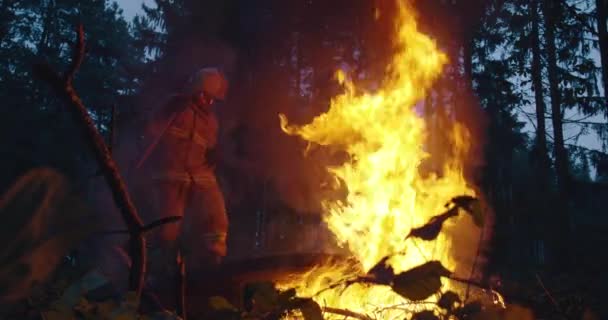 Bombero con equipo de seguridad y hacha extinguiendo fuego en el bosque por la noche — Vídeos de Stock