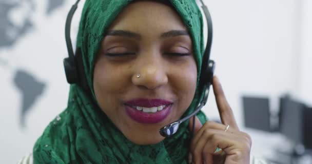 Αφρικανική ισλαμίστρια επιχειρηματίας με μαντίλα φορώντας ακουστικά ενώ στεκόταν στο σύγχρονο γραφείο — Αρχείο Βίντεο