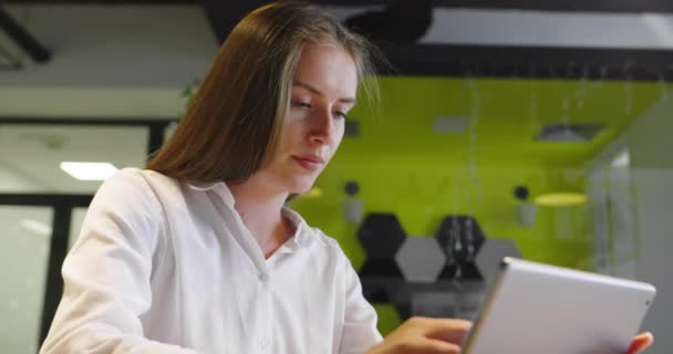 Красивая молодая девушка с длинными волосами, держащая цифровой планшет чтение электронной почты сидя на диване в офисе стартапа — стоковое видео