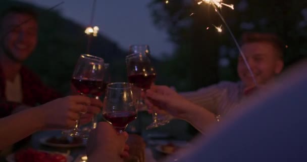 スプリンクラーを使用して休暇を祝う幸せな友人のグループと赤いワインを飲みながら 川の近くでピクニックフランスのディナーパーティーを屋外で開催 — ストック動画