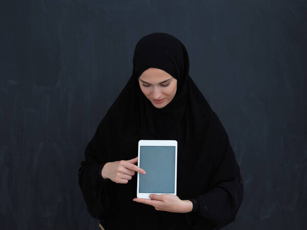 Young Arab businesswoman in traditional clothes or abaya and glasses showing tablet computer display in front of black chalkboard representing islam fashion and technology